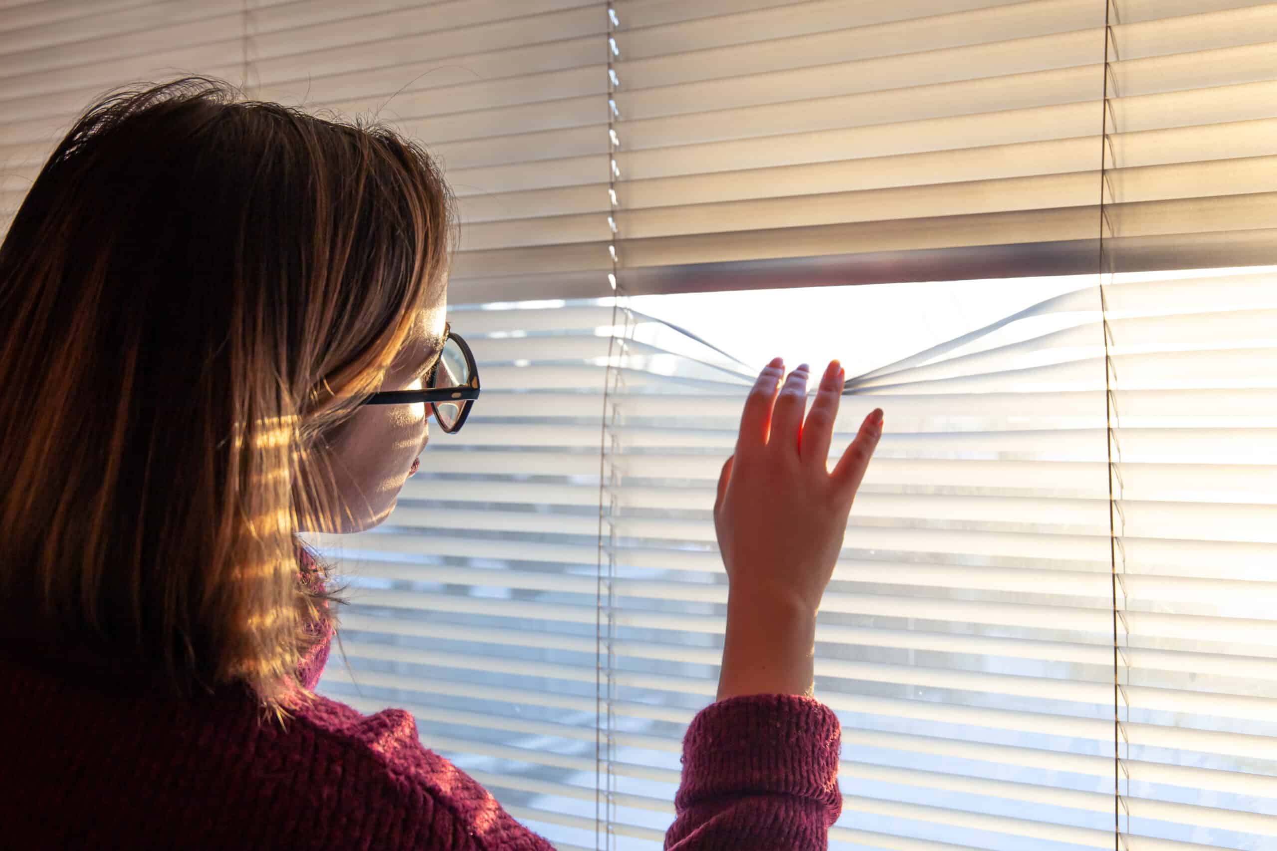 A woman with glasses looks through the blinds at the early morning sunlight. Grow on plants.